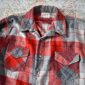 May include: A vintage red, grey, and black plaid flannel shirt with two chest pockets. The shirt has a collared neckline and button-down front. The fabric appears to be a thick, warm material, suitable for cooler weather.