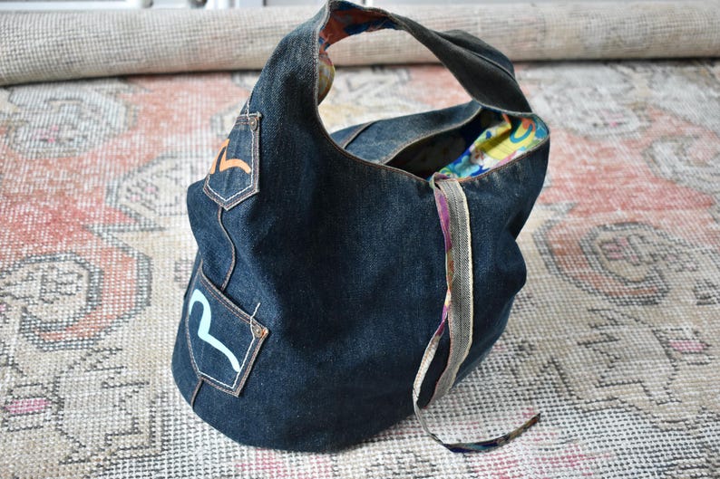 Puede incluir: Bolso hobo de mezclilla azul oscuro con una sola correa para el hombro. El bolso presenta dos peque&ntilde;os bolsillos de mezclilla con dise&ntilde;os coloridos. El forro interior es un vibrante estampado floral. Un accesorio elegante y pr&aacute;ctico.