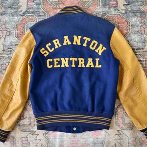 Può includere: Giacca college vintage con corpo blu navy e maniche in pelle marrone chiaro. La schiena presenta la scritta "SCRANTON CENTRAL" in lettere gialle. La giacca ha colletto, polsini e orlo a coste blu navy e giallo a righe.