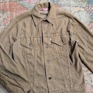 May include: A tan corduroy jacket with a button-up front, two chest pockets, and two side pockets. The jacket has a collar and long sleeves. The Levi's brand tag is visible on the collar.