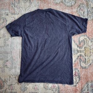May include: A solid dark charcoal gray short-sleeve t-shirt with a crew neckline. The garment is displayed flat, showing the back side of the shirt, which features a plain, unadorned fabric surface with no visible graphics, text, or patterns.