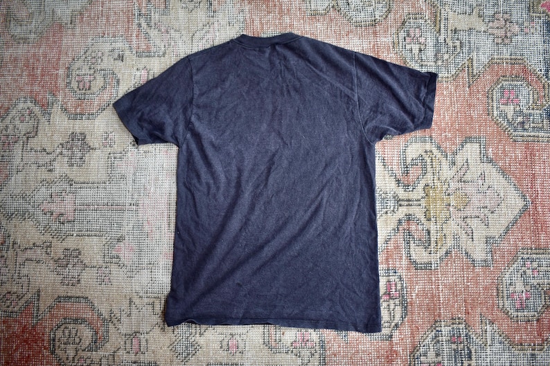 May include: A solid dark charcoal gray short-sleeve t-shirt with a crew neckline. The garment is displayed flat, showing the back side of the shirt, which features a plain, unadorned fabric surface with no visible graphics, text, or patterns.