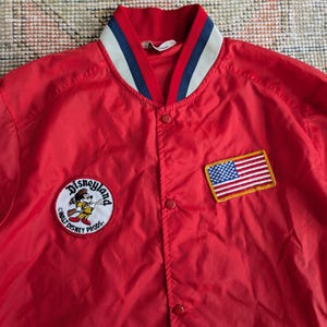 May include: Red satin baseball jacket with a white and blue striped collar. The jacket features a Mickey Mouse logo with the text "Disneyland" and "Walt Disney Prod.". The jacket also has a small American flag patch.