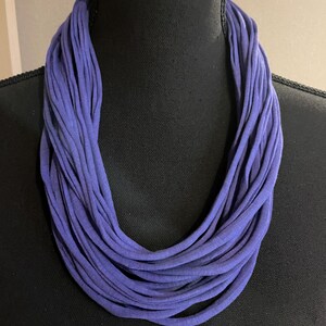 Op de afbeelding: Een gelaagde infinity sjaal in een levendige paarse tint. De sjaal is gemaakt van zachte stof en is gedrapeerd rond een zwarte mannequin. Dit accessoire voegt een vleugje kleur en textuur toe aan elke outfit.