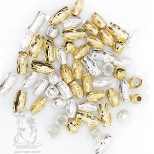 May include: A collection of gold and silver metal beads with an engraved design. The beads are oval shaped and have a hole in the center for stringing.