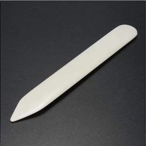 May include: A white bone folder for scoring and creasing paper. It is a long, thin tool with a pointed tip and a rounded end.