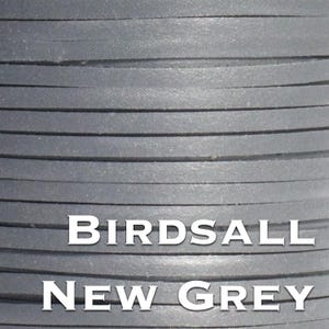 Op de afbeelding: Close-up van een spoel met platte, grijze leren strips. Het leer heeft een effen, neutrale kleur en de textuur lijkt glad. De tekst "BIRDSALL NEW GREY" is in het wit gedrukt.