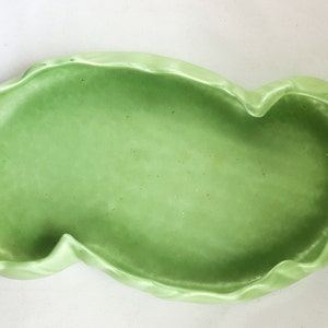 Puede incluir: Una bandeja de cerámica verde claro con una forma orgánica única. La bandeja tiene un borde ondulado y festoneado y un acabado liso y brillante. El diseño general recuerda a una hoja o una forma natural, adecuada para servir alimentos o como pieza decorativa.