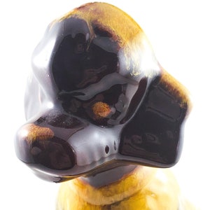 May include: A ceramic figurine of a brown and yellow poodle dog. The dog is sitting with its head tilted to the side. The figurine is glazed with a brown and yellow glaze.