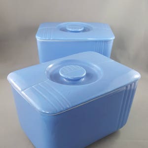 Könnte beinhalten: Zwei blaue Keramik-Aufbewahrungsbehälter mit Deckeln. Die Behälter sind quadratisch mit abgerundeten Ecken und haben ein erhabenes Design auf dem Deckel.