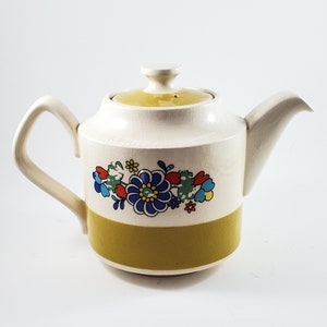 May include: A white ceramic teapot with a yellow lid and a floral design in shades of blue, red, green, and yellow. The teapot has a wide, round body and a spout on the right side.