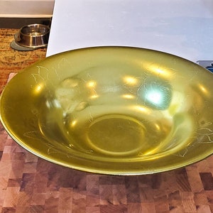 May include: A large, gold-coloured metal bowl with a decorative etched design. The bowl is sitting on a wooden chopping board.