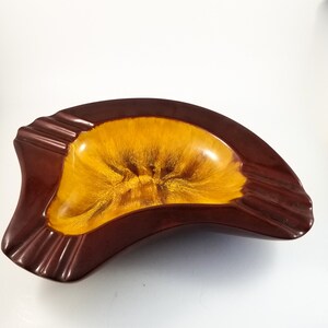 May include: A brown and yellow ceramic ashtray shaped like a crescent moon. The ashtray has a deep well in the center and a raised rim around the edge.
