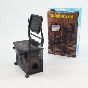 May include: A vintage metal pencil sharpener shaped like a vanity with a mirror. The sharpener is brown and has a drawer and a small hole for sharpening pencils. The box is blue and white and says "Washstand" on it.