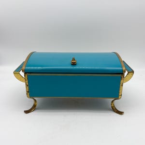 Peut inclure: Une boîte rectangulaire turquoise avec un couvercle incurvé. Elle est ornée de bordures et de pieds dorés. Un petit bouton doré orne le dessus. La boîte est probablement vintage ou antique.