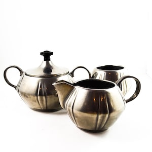 以下が含まれることがあります： 蓋付きの砂糖入れ、クリーマー、砂糖入れの3点セットの銀色のティーセット。セットはフルーテッドデザインで、蓋には黒いノブが付いています。