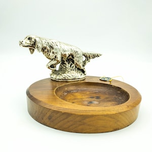 May include: A silver-colored figurine of a dog, possibly a pointer, standing on a wooden base with a small round dish. The dog is in a running pose with its head turned to the side.