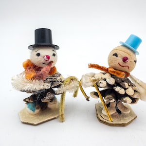 May include: Two handmade snowman figurines. One snowman wears a black top hat and the other a blue top hat. Both snowmen have a pinecone body, a felt base, and a pipe cleaner cane. They have orange scarves and red noses.