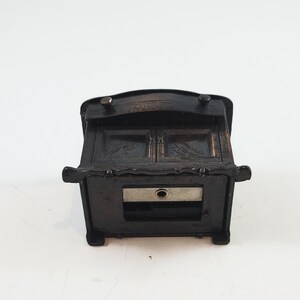 May include: A black metal pencil sharpener shaped like a small cabinet with a drawer. The drawer is open and reveals a metal blade.