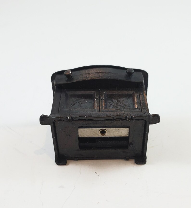 May include: A black metal pencil sharpener shaped like a small cabinet with a drawer. The drawer is open and reveals a metal blade.