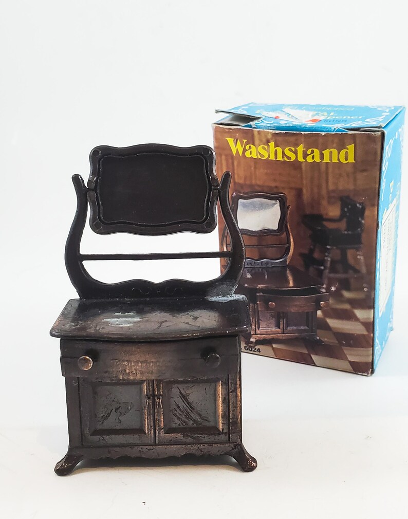 May include: A vintage metal pencil sharpener shaped like a washstand with a mirror and a drawer. The sharpener is brown and has a woodgrain finish. The box says "Washstand" on it.