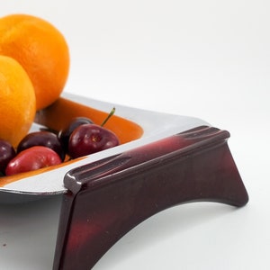 May include: A white and orange ceramic fruit bowl with a dark red stand. The bowl is filled with oranges and cherries.