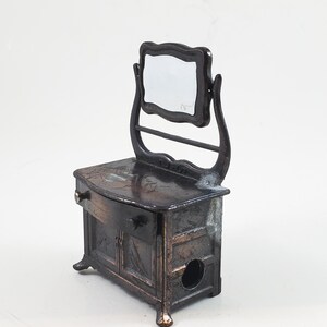 May include: A vintage metal pencil sharpener shaped like a vanity with a mirror. The sharpener is painted in a dark bronze colour and has a drawer and two doors. The mirror is attached to the top of the vanity and has a simple, ornate frame.