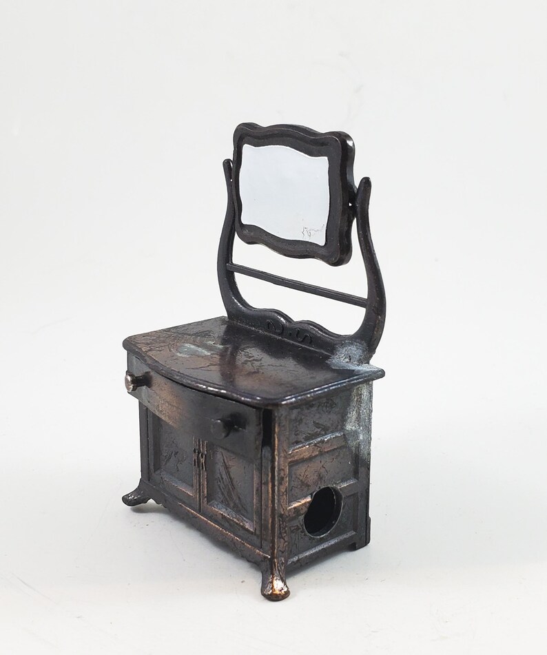 May include: A vintage metal pencil sharpener shaped like a vanity with a mirror. The sharpener is painted in a dark bronze colour and has a drawer and two doors. The mirror is attached to the top of the vanity and has a simple, ornate frame.