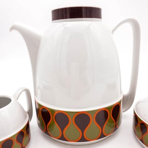 May include: A white ceramic coffee pot with a brown, green, and orange geometric pattern. The pot has a spout and a handle. There are two matching creamer and sugar bowls.