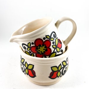 May include: A white ceramic creamer and sugar bowl set with a floral design. The design features red, green, and black flowers on a white background.