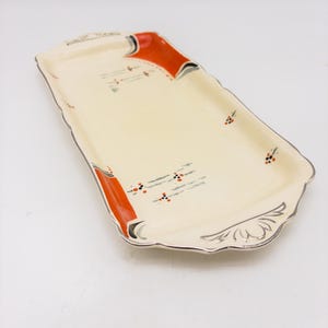 May include: A rectangular ceramic serving tray with a cream-coloured base, featuring orange and black geometric accents at the corners and small floral motifs. The edges are scalloped and finished with a silver-toned metallic trim.