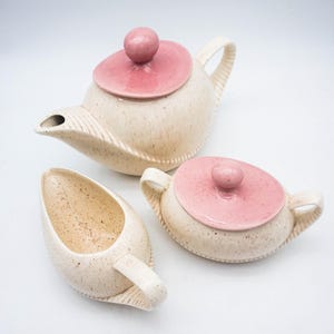 May include: A vintage ceramic tea set comprising a teapot, sugar bowl, and creamer. The set has a speckled beige finish with pink lids and accents. The teapot has a long spout and a curved handle. The sugar bowl has two handles.