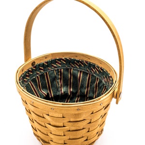 May include: A woven basket with a wooden handle and a green and brown striped fabric lining. The basket is empty and is sitting on a white background.