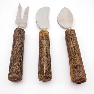 May include: Set of three stainless steel cheese knives with wooden handles. The handles are a dark brown color and have a wood grain pattern. The knives have different blade shapes, including a fork, a curved blade, and a pointed blade.