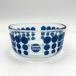 May include: Clear glass Pyrex bowl with a blue polka dot pattern. The bowl has a rounded shape and features the Pyrex logo and "1915" printed in blue on the front. A vintage kitchenware item.