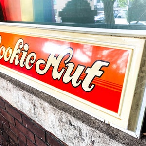 May include: A vintage-style sign with a white frame and orange background. The sign reads "Cookie Hut" in a stylized font.