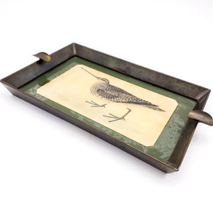 Könnte beinhalten: Ein rechteckiges Tablett aus Metall mit einer grünen und goldenen Oberfläche. Das Tablett zeigt eine Schwarzweißillustration eines Vogels mit langen Beinen und einem langen Schnabel. Das Tablett hat eine kleine, gebogene Vertiefung auf einer Seite, wahrscheinlich als Aschenbecher.