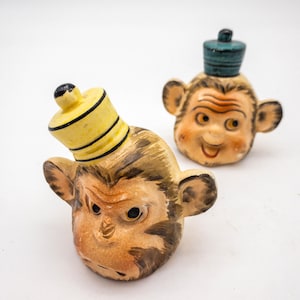 May include: Two ceramic monkey head figurines with painted details. One monkey is wearing a yellow and black striped hat, and the other is wearing a blue hat.
