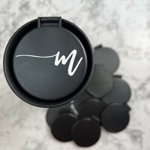 May include: A black compact mirror with a white "M" on the lid. The mirror is surrounded by several other black compact mirrors.