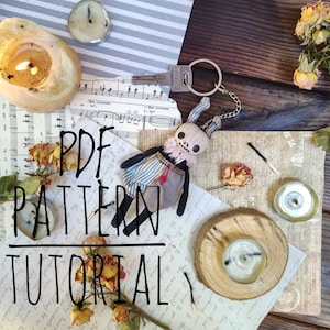 Puede incluir: Un patrón PDF y un tutorial para hacer una pequeña muñeca hecha a mano con una cara de calavera, extremidades negras y un vestido a rayas. La muñeca lleva un llavero y está rodeada de objetos decorativos, como velas, flores secas y partituras.