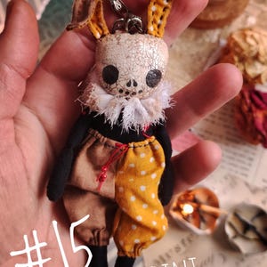 May include: A handmade doll with a skull-like face, black eyes, and a white ruffled collar. The doll wears a black top, brown trousers, and yellow polka-dot trousers. The text "#15 ORANGE ON POINT" is visible.