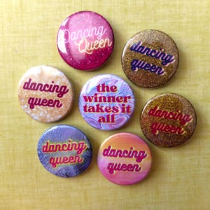 Könnte beinhalten: Sieben runde Ansteckbuttons mit Glitzer- und Marmoreffekten. Die Buttons zeigen die Sätze "Dancing Queen" und "The Winner Takes It All" in Pink, Gold und Blau. Die Buttons sind auf einer gelb-grünen Oberfläche angeordnet. Ein lustiges Accessoire.