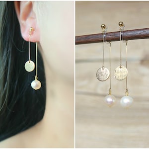 May include: Gold-toned earrings featuring a textured disc and a dangling white pearl. The earrings are long and slender, with a simple design. The earrings are shown on a person's ear and also displayed on a wooden rod.
