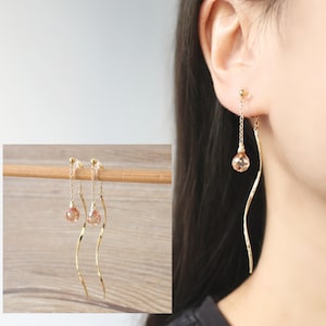 May include: A pair of gold dangle earrings with a small, teardrop-shaped crystal at the end of each earring. The earrings are shown on a wooden surface and on a model's ear.