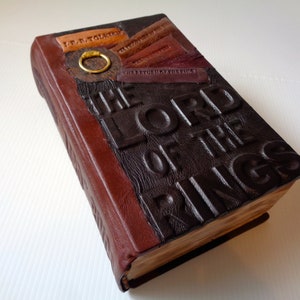 Könnte beinhalten: Ein braunes, ledergebundenes Buch mit dem Titel "Der Herr der Ringe" auf dem Einband geprägt. Das Buch hat einen goldenen Ring auf dem vorderen Einband.