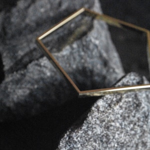 Könnte beinhalten: Ein goldener sechseckiger Rahmen liegt auf zwei grauen Steinen. Der Rahmen besteht aus dünnem Metall und hat eine glatte, polierte Oberfläche.