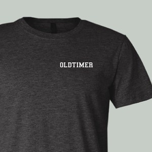 May include: A dark heather grey t-shirt with the word "OLDTIMER" printed in white on the front.