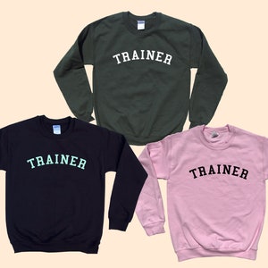 Pu&ograve; includere: Tre felpe girocollo in diversi colori: verde scuro, blu navy e rosa. Ogni felpa ha la scritta "TRAINER" stampata sul petto in un colore a contrasto. Le felpe sono esposte su uno sfondo beige chiaro.