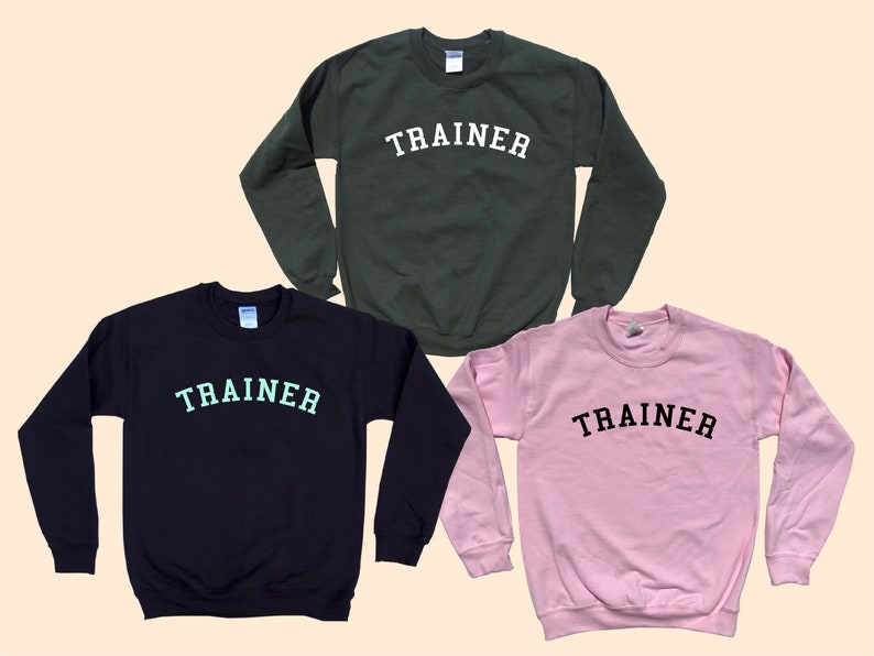 Pu&ograve; includere: Tre felpe girocollo in diversi colori: verde scuro, blu navy e rosa. Ogni felpa ha la scritta "TRAINER" stampata sul petto in un colore a contrasto. Le felpe sono esposte su uno sfondo beige chiaro.