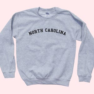 May include: A light gray sweatshirt with the text "NORTH CAROLINA" arched across the chest in black letters. The long-sleeved crew neck top is made of a soft material. The sweatshirt is a casual style.